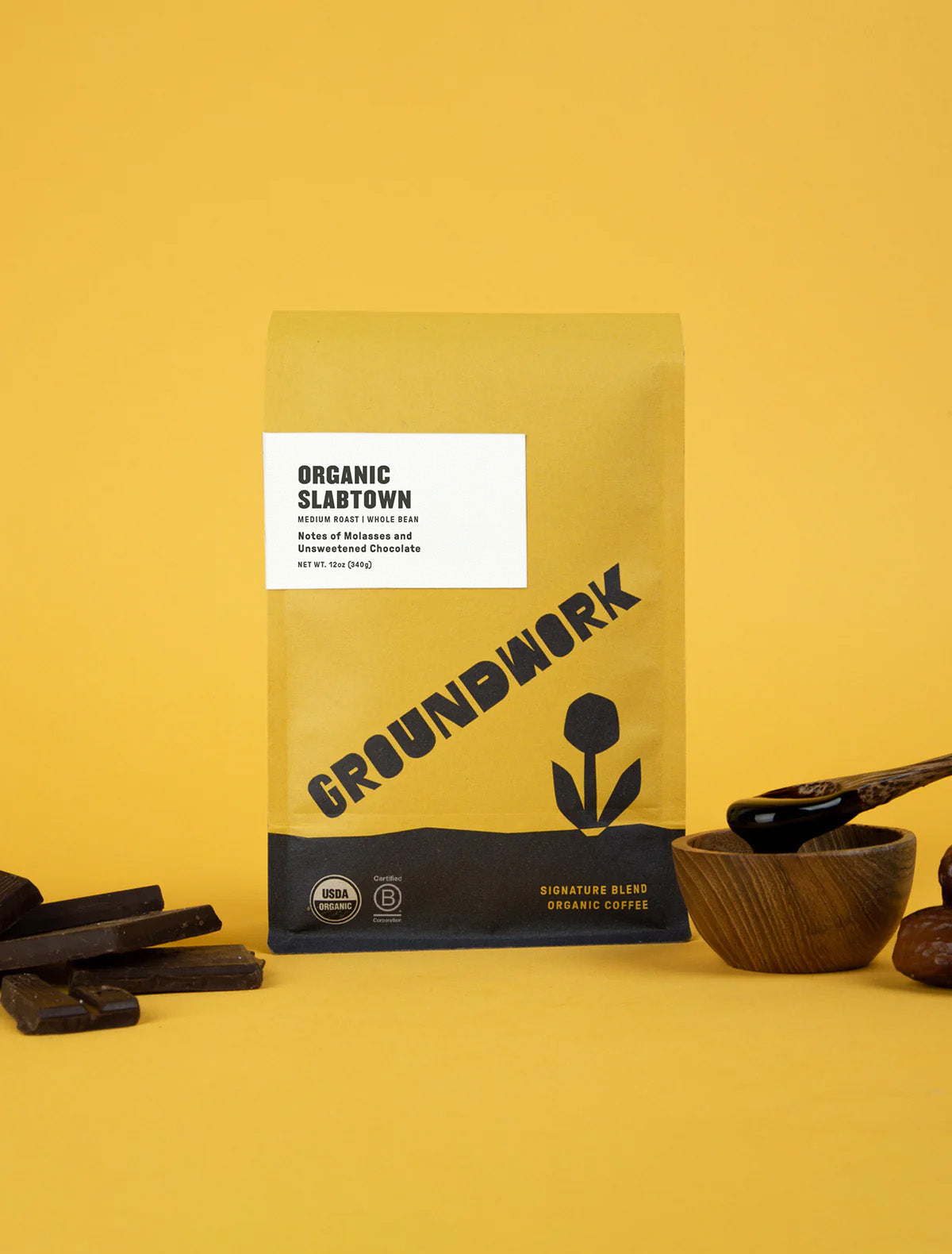 A yellow bag of Groundwork Coffee Slabtown sits on a yellow background, next to pieces of unsweetened chocolate, a wooden bowl, and a wooden spoon.