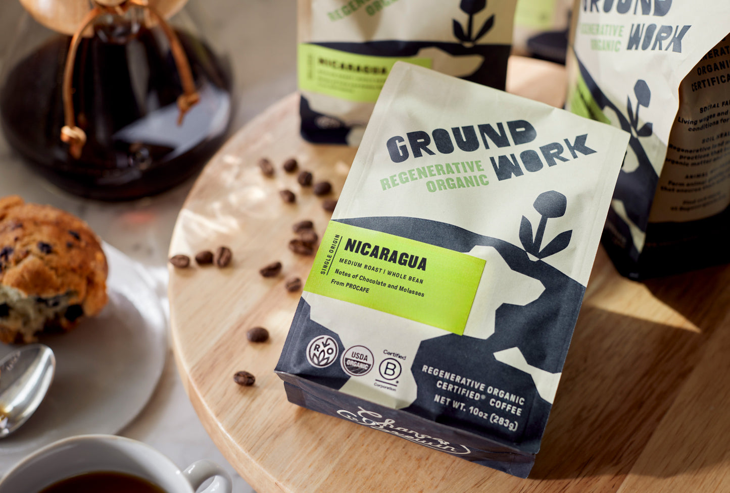 Groundwork Regenerative Organic Certified® Nicaragua coffee bag on a wooden table with scattered coffee beans, a muffin, a cup of coffee, and a Chemex brewer in the background. Medium roast whole bean, single origin from PROCAFE with tasting notes of chocolate and molasses. Certified USDA Organic, Regenerative Organic Certified®, and B Corporation. Net weight 10 oz (283 g).