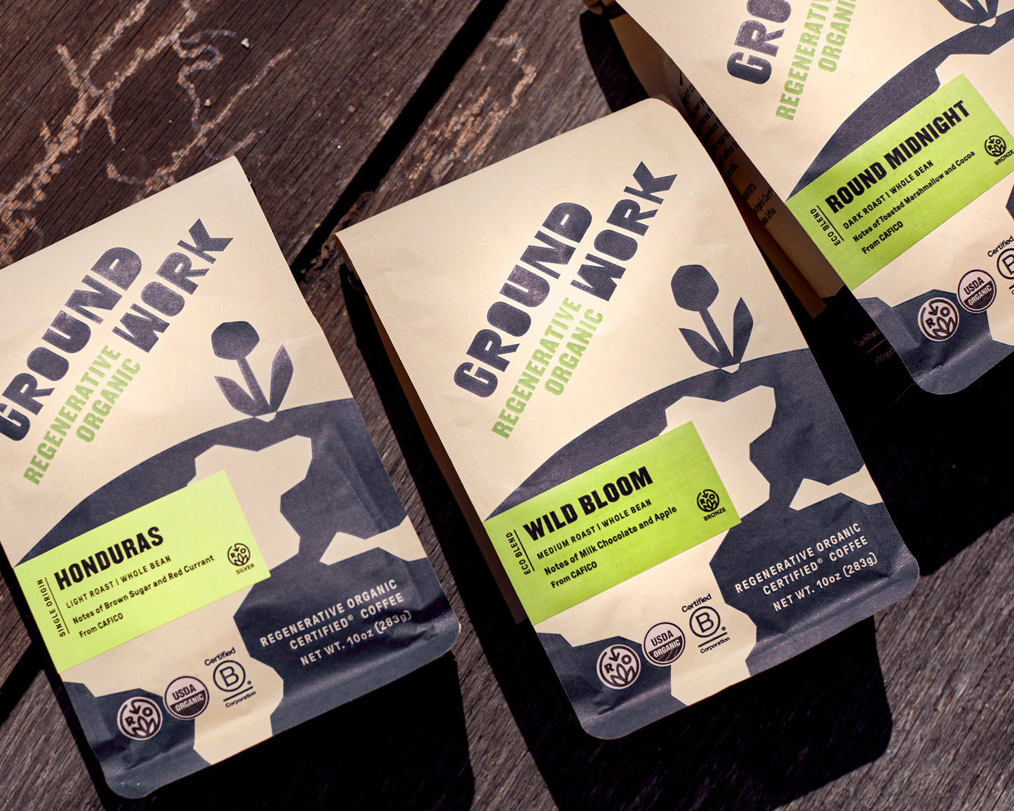 Three bags of Groundwork Coffee’s Regenerative Organic Certified® Honduras CAFICO sit on a sunlit wood surface, featuring earthy packaging and green labels.