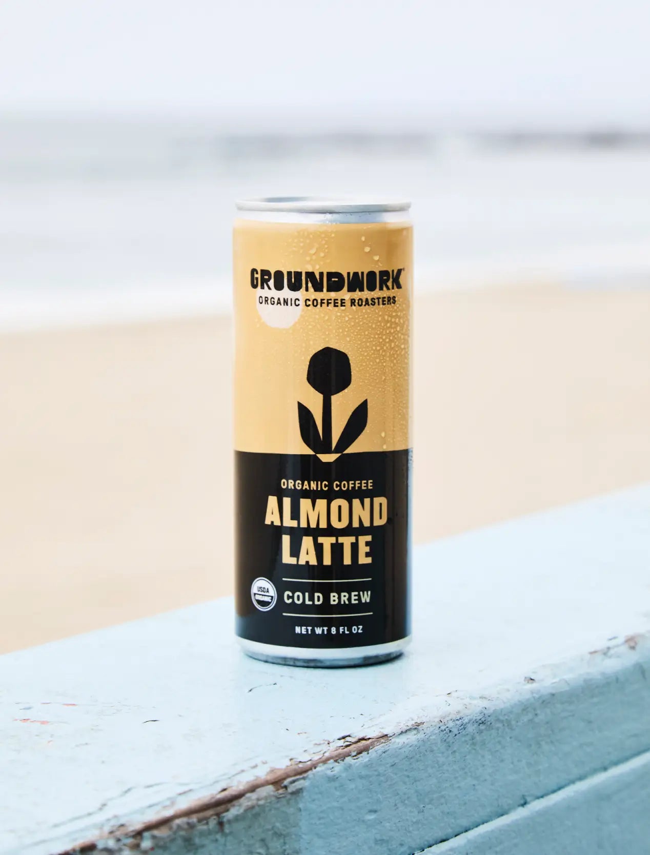 A can of Groundwork Coffee Organic Cold Brew Latte rests on a light blue wooden surface with a blurred beach in the background.