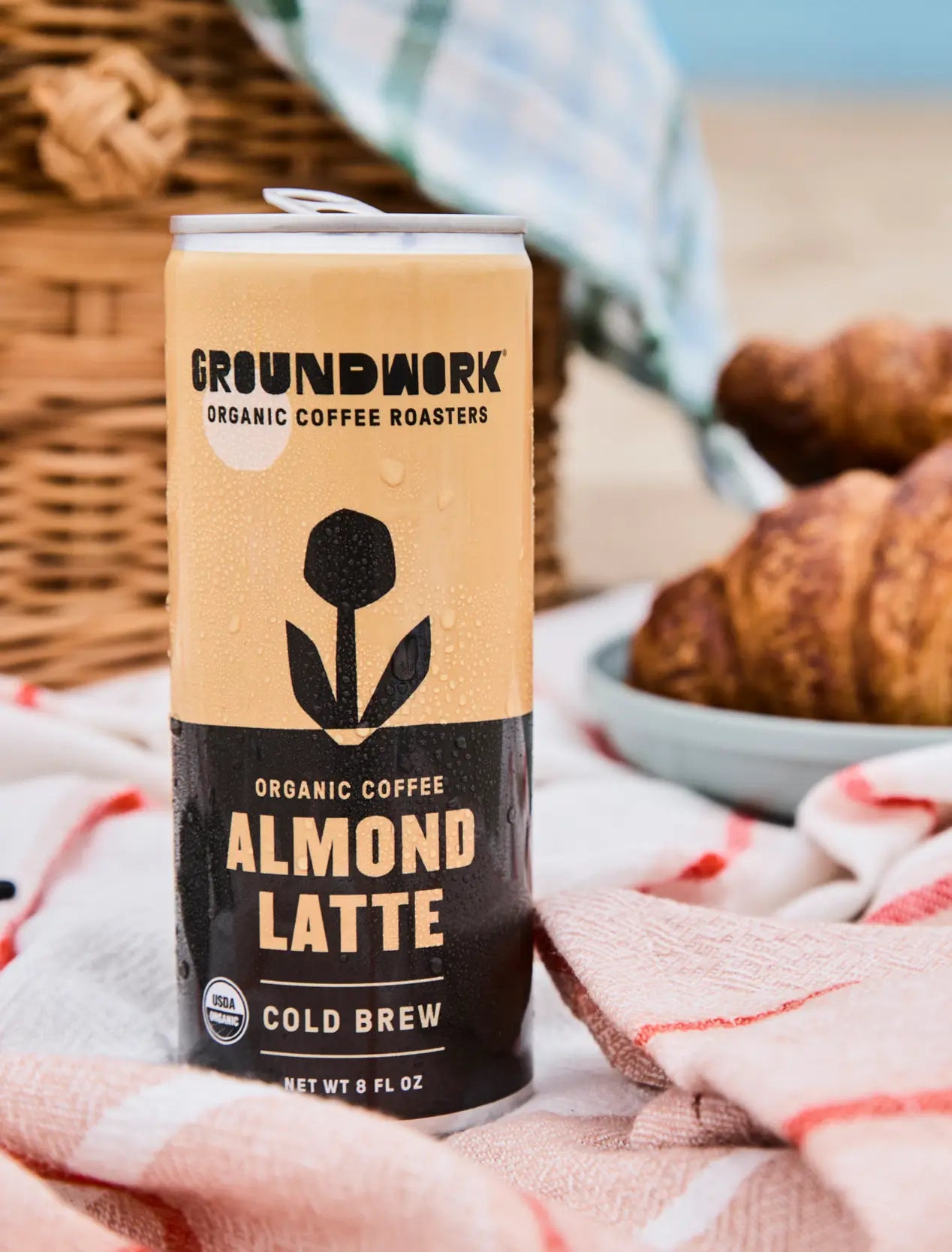 A can of Groundwork Coffee Organic Cold Brew Latte rests on a picnic blanket with croissants and a wicker basket in the background.