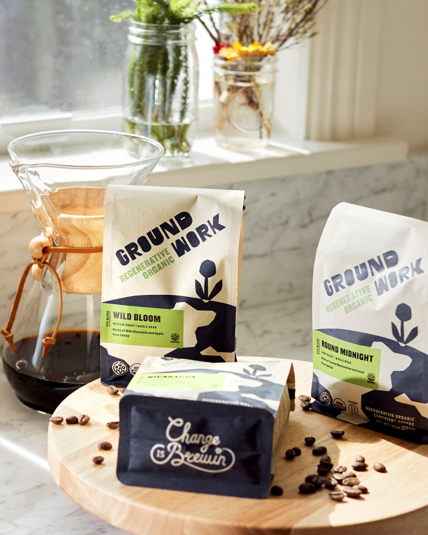 A bag of Groundwork Coffee’s Regenerative Organic Certified® Round Midnight sits on a wooden board with cocoa-hued beans and a Chemex by a sunny window.