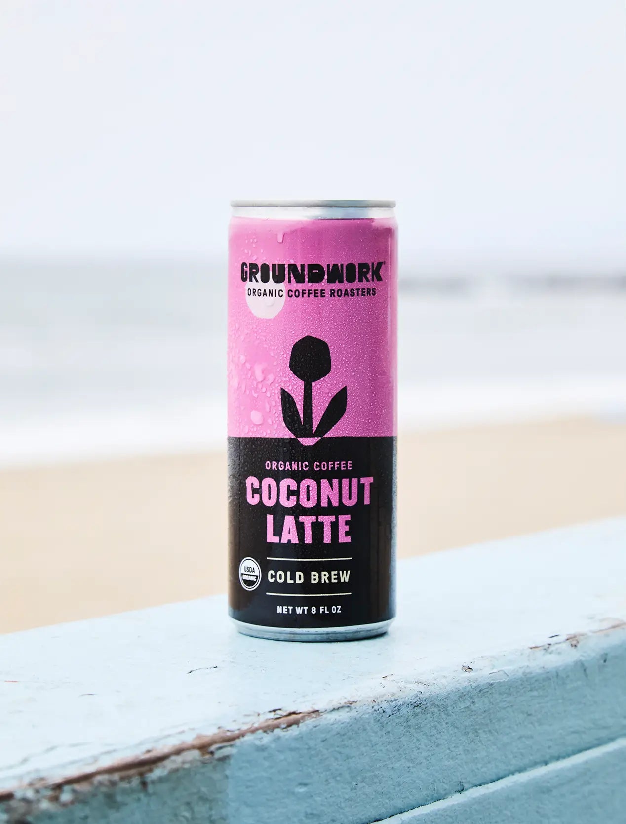 A can of Groundwork Coffee Organic Cold Brew Latte rests on a blue wooden surface with a blurred beach and ocean in the background.