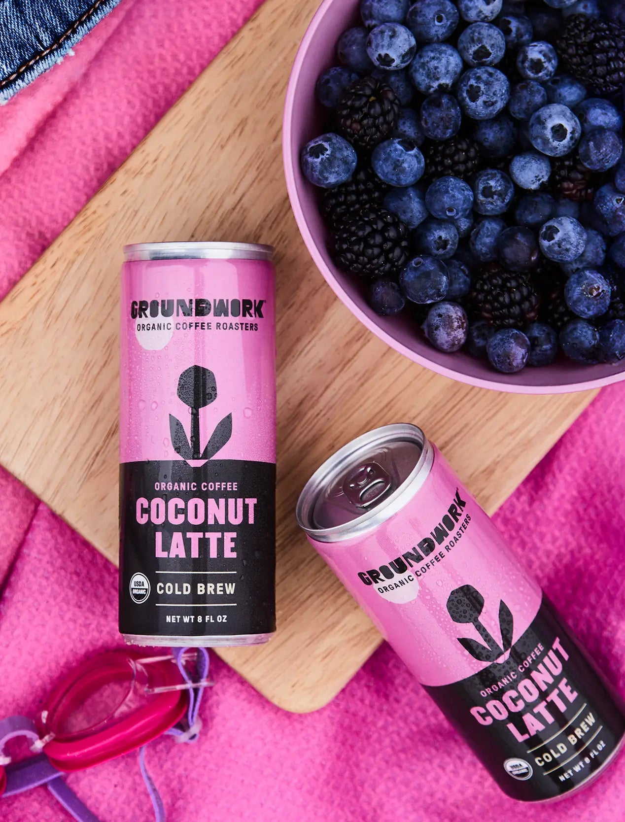 Two cans of Groundwork Coffee Organic Cold Brew Latte on a wooden board with blueberries, blackberries, and pink sunglasses on a pink cloth.