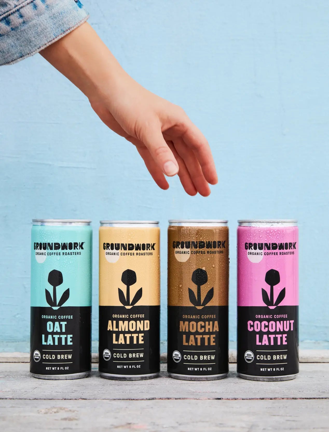 A hand reaches for four cans of Groundwork Coffee Organic Cold Brew Latte—Oat, Almond, Mocha, and Coconut—lined up against a blue background.
