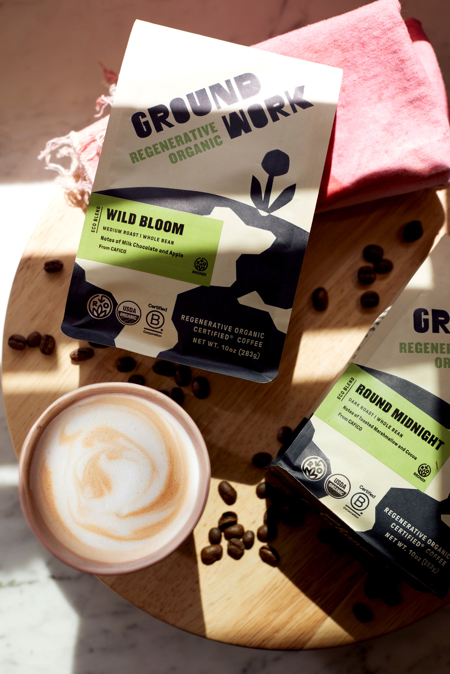 Two bags of Groundwork Coffees Regenerative Organic Certified® Round Midnight on a wooden board with coffee beans and a latte, featuring cocoa and toasted marshmallow notes.