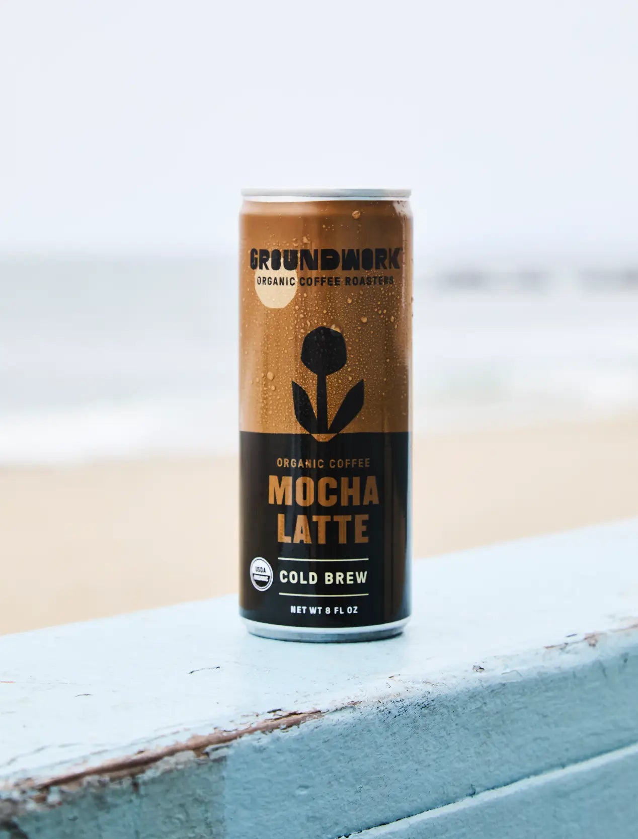 A can of Groundwork Coffee Organic Cold Brew Latte rests on a blue wooden surface with a blurred beach and ocean in the background.