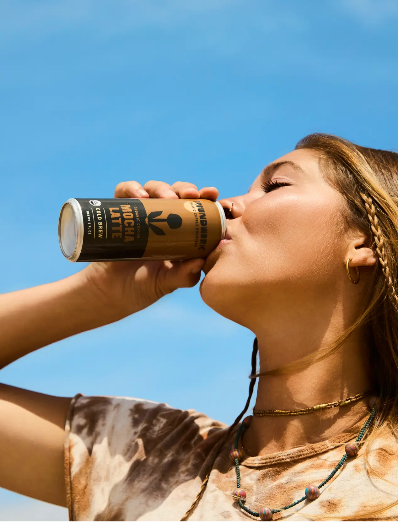 A woman with braided hair sips Groundwork Coffee’s Organic Cold Brew Latte from a can, wearing a tie-dye shirt and layered necklaces under a clear blue sky.