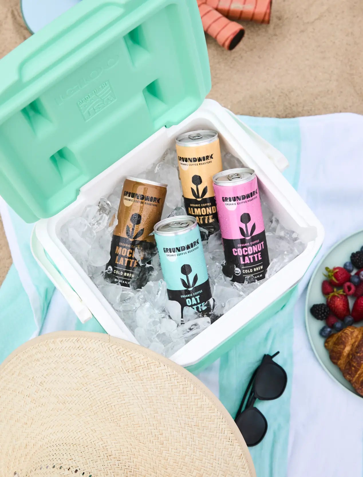 A cooler with ice holds four cans of Groundwork Coffee’s Organic Cold Brew Latte, set on a striped beach towel with a sunhat, sunglasses, and a plate of pastries, berries, and grapes.