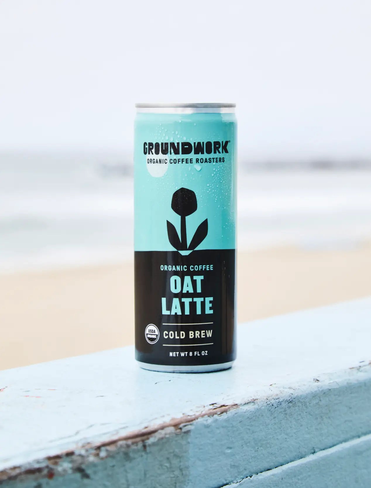 A can of Groundwork Coffee Organic Cold Brew Latte rests on a light blue wooden surface with a blurry beach background.