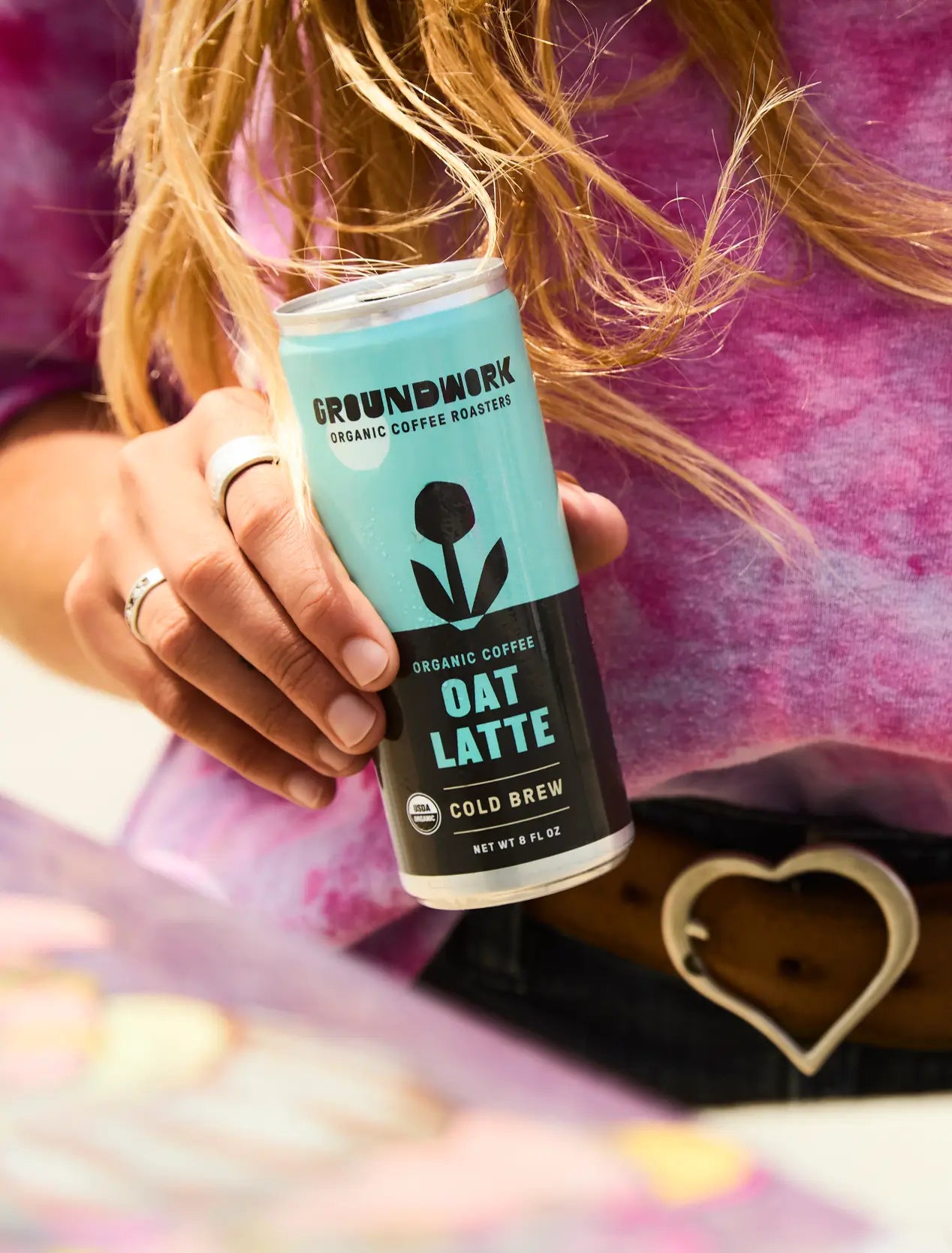 A person in a pink tie-dye shirt with a heart-shaped belt buckle holds a can of Groundwork Coffee Organic Cold Brew Latte.