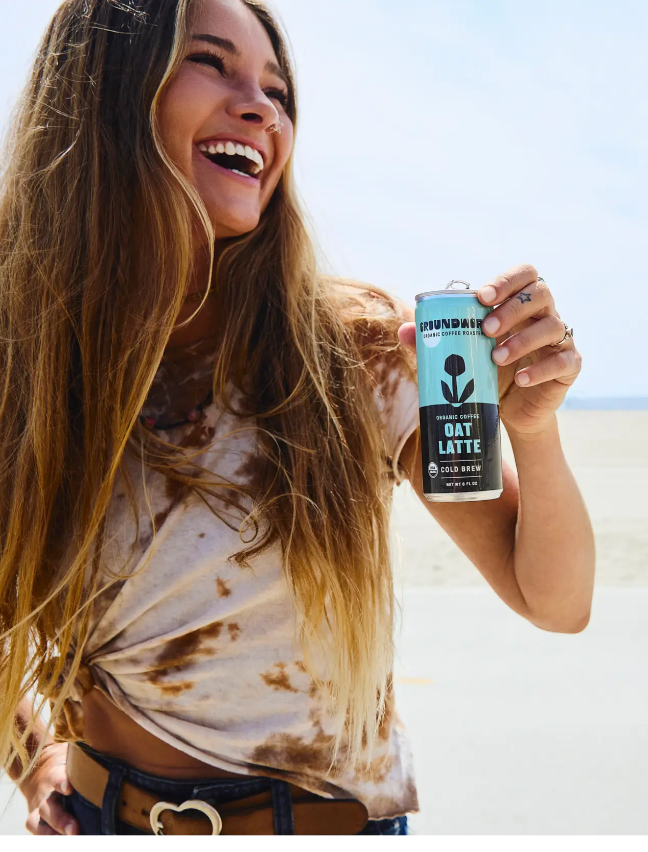 A smiling woman with long hair holds a can of Groundwork Coffee Organic Cold Brew Latte while standing outdoors on a sunny day.