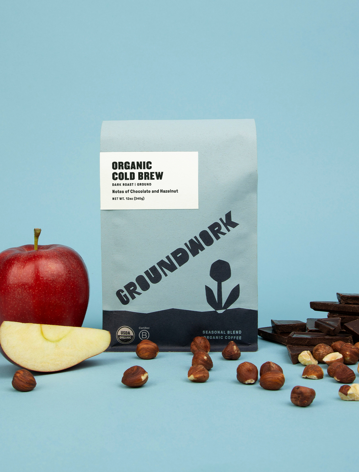 A bag of Groundwork Coffee Stores Cold Brew Blend sits with apple slices, hazelnuts, and dark chocolate on a light blue background.