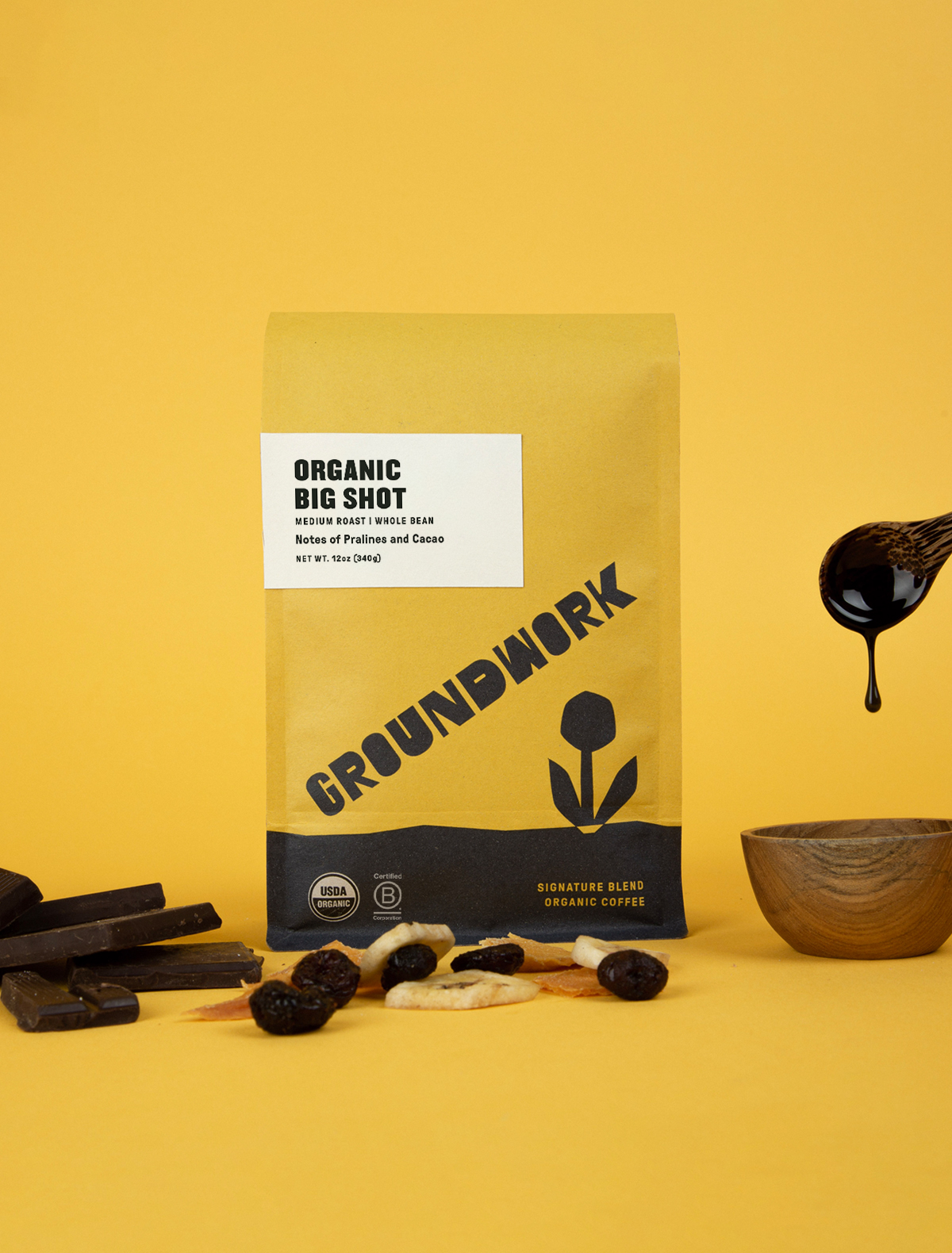 A yellow bag of Groundwork Coffee’s Big Shot stands on a yellow background, surrounded by chocolate, pralines, dried fruits, and a spoon dripping with dark liquid.