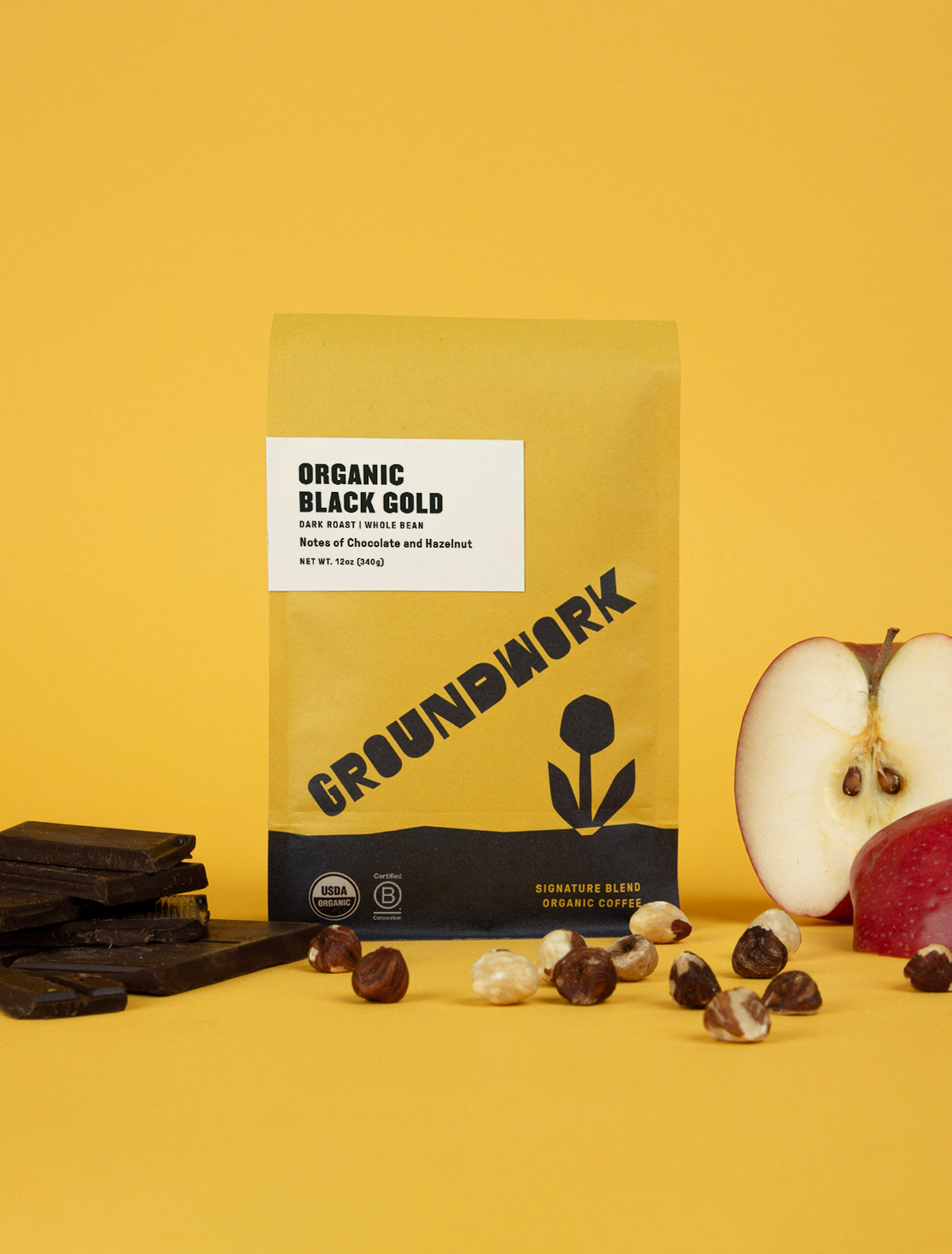 A bag of Groundwork Coffee Co Black Gold stands on a yellow background, surrounded by chocolate pieces, hazelnuts, and a halved red apple.