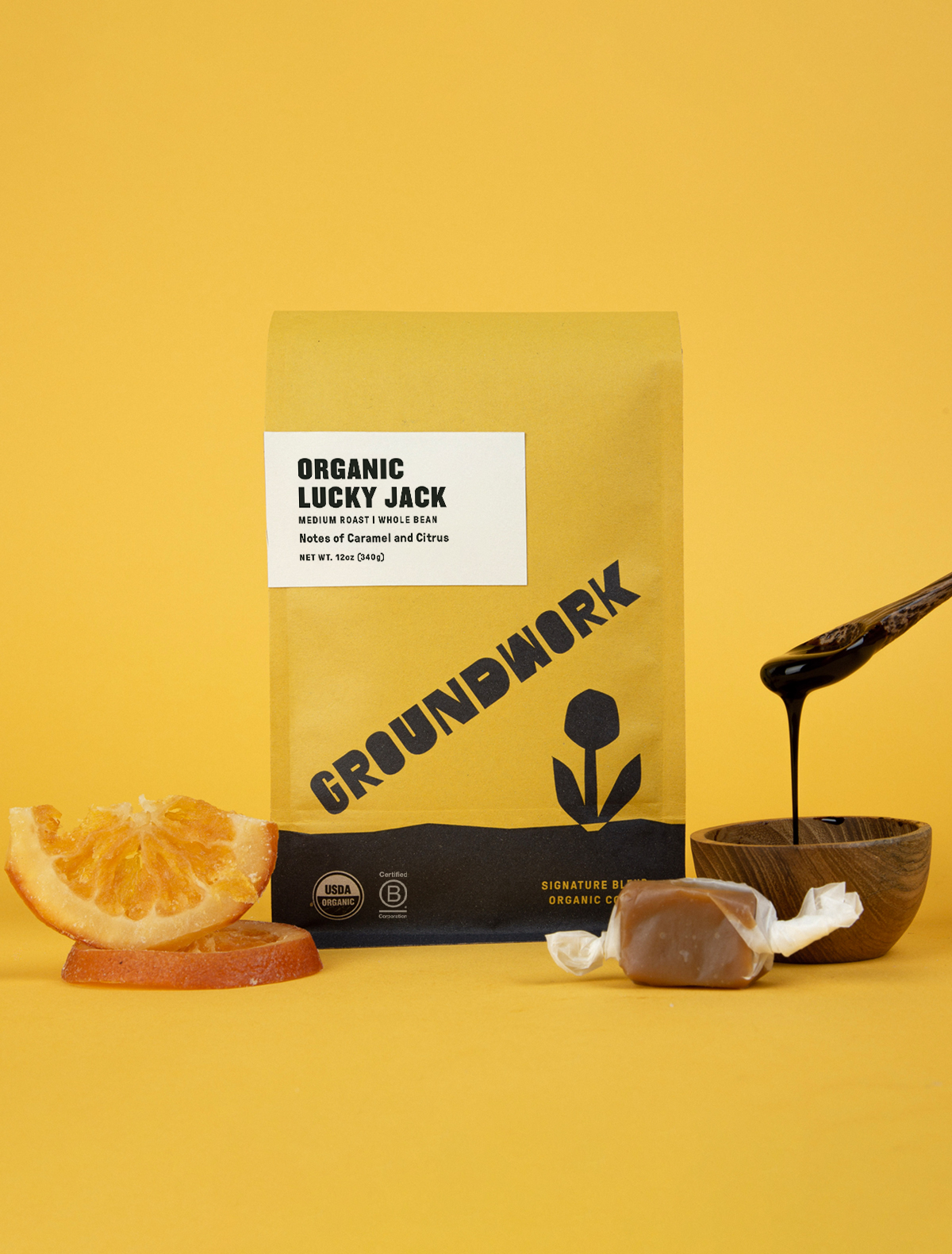 A yellow bag of Groundwork Coffees Lucky Jack stands on a yellow surface, with orange slices, a caramel, and a spoon dripping caramel beside it.