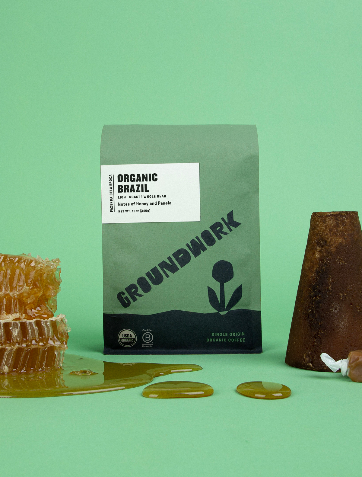 A green bag of Groundwork Coffee Stores Brazil: Fazenda Bela Epoca stands on a green surface beside honeycomb, honey, and an upside-down coffee cup.