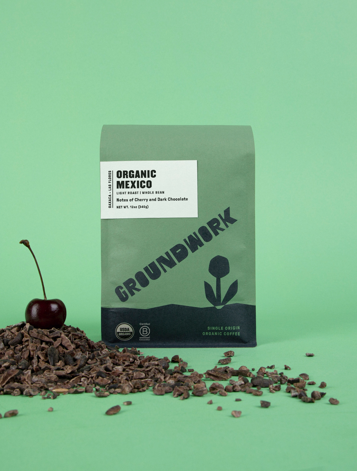 A green bag of Groundwork Coffee Store’s Mexico: Oaxaca - Las Flores light roast sits on a green surface with cocoa nibs and a cherry, set against a green background.