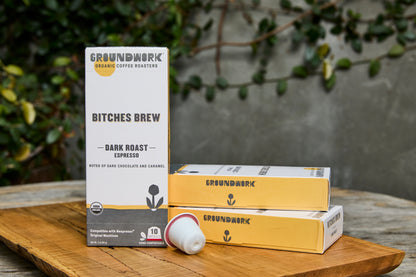 A box and two sleeves of Groundwork Coffee Organic Bitches Brew Nespresso pods sit on a wooden surface with greenery in the background.