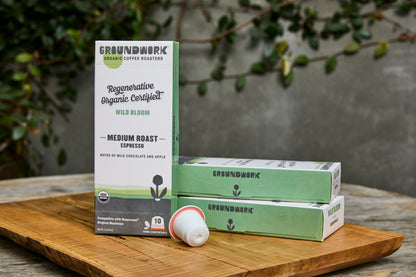 A box of Groundwork Coffee Regenerative Organic Certified® Wild Bloom - Nespresso pods, with two boxes and one pod displayed on a wooden board against leafy greenery.