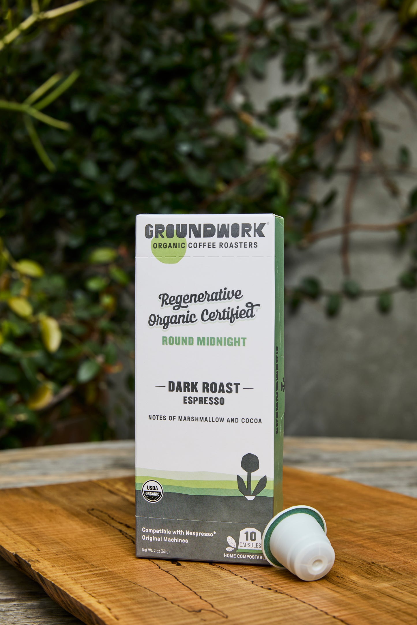 A box of Groundwork Coffee Regenerative Organic Certified® Round Midnight - Nespresso capsules sits on a wooden table with plants and a blurred garden background.