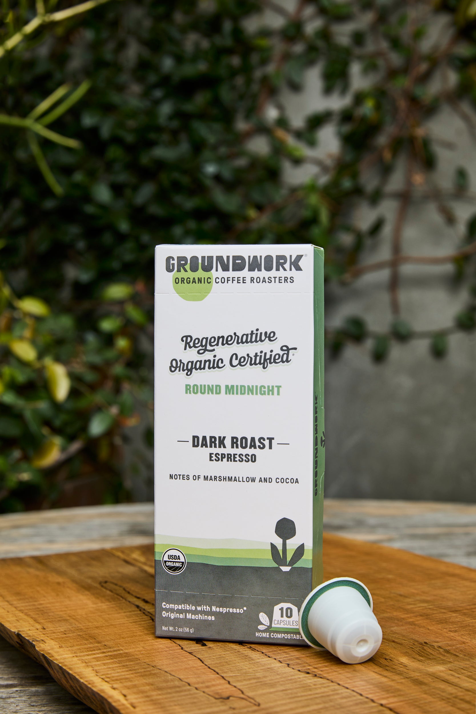 A box of Groundwork Coffee Regenerative Organic Certified® Round Midnight - Nespresso capsules sits on a wooden table with plants and a blurred garden background.
