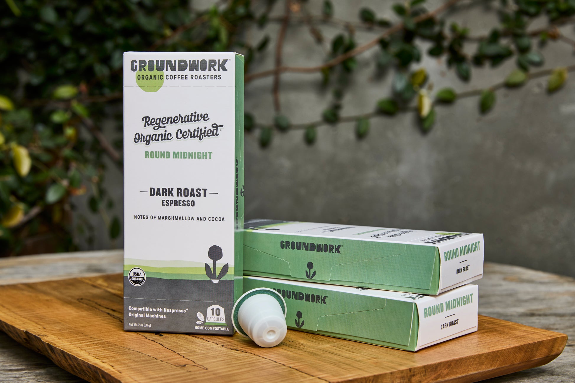 Boxes of Groundwork Coffee’s Regenerative Organic Certified® Round Midnight - Nespresso capsules are displayed on a wooden board with green foliage and a gray wall in the background.