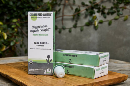 Boxes of Groundwork Coffee’s Regenerative Organic Certified® Round Midnight - Nespresso capsules are displayed on a wooden board with green foliage and a gray wall in the background.