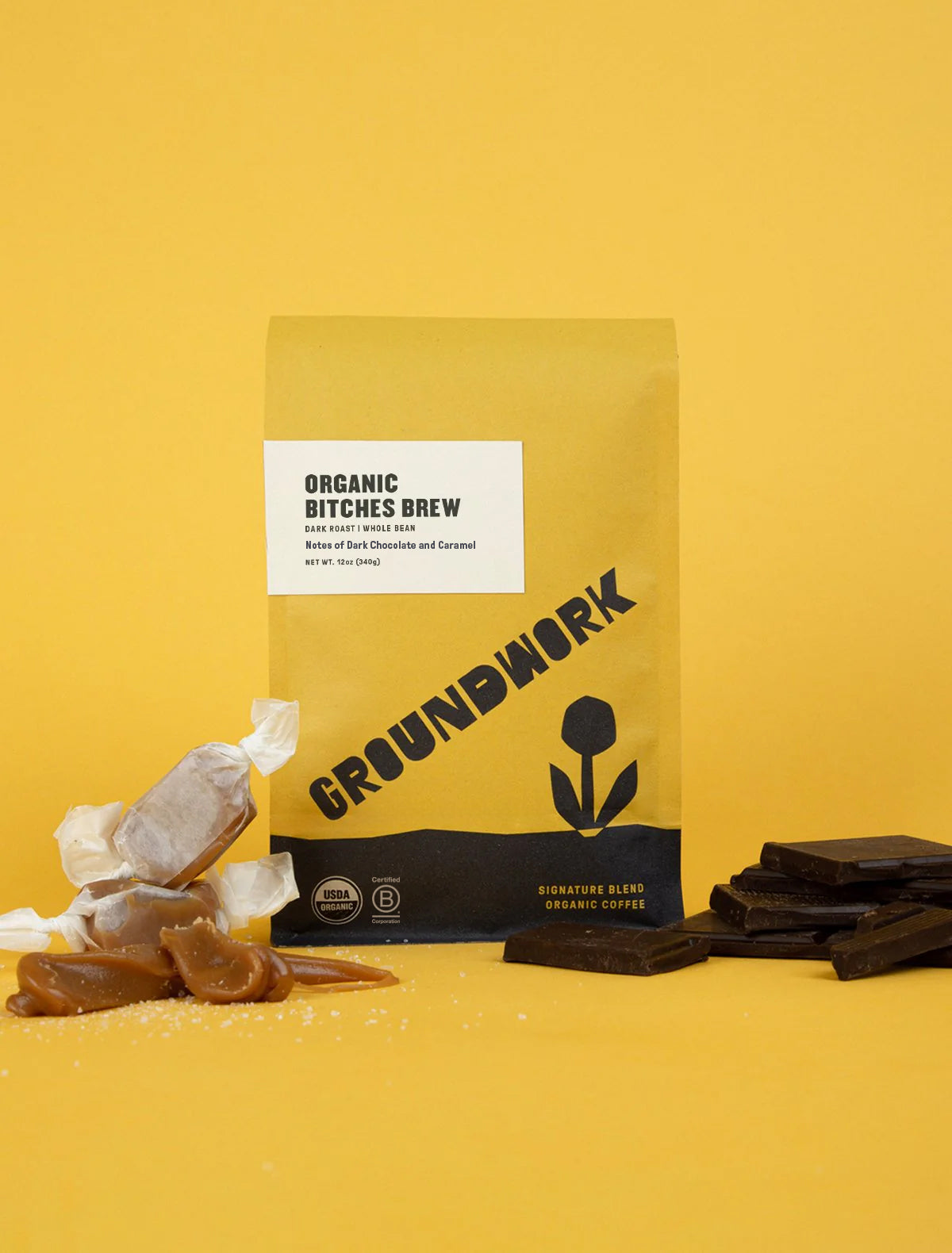 A yellow bag of Groundwork Coffee Store's Bitches Brew sits on a yellow surface with caramel and dark chocolate beside it.
