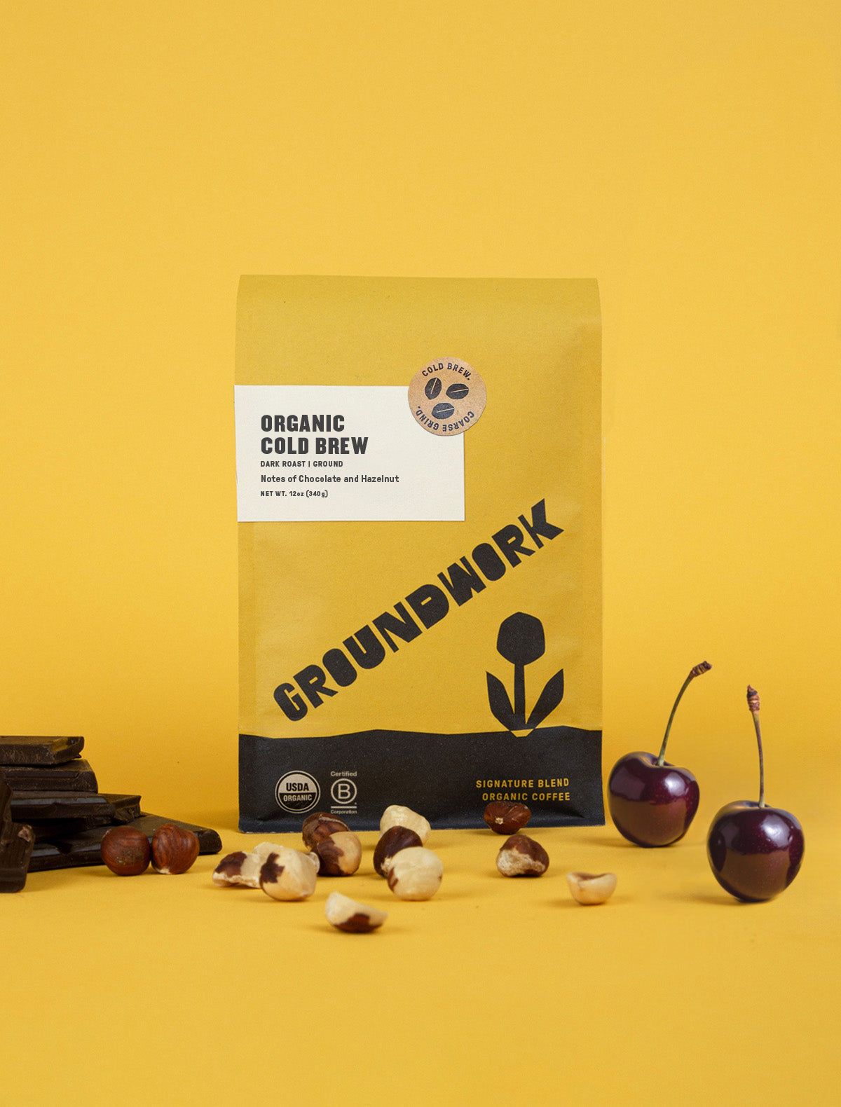 A yellow bag of Groundwork Coffee Store's Organic Cold Brew Blend stands on a yellow background, surrounded by coffee beans, hazelnuts, and two cherries.