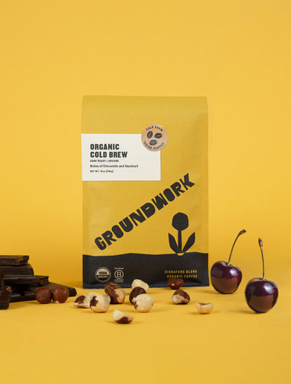 A yellow bag of Groundwork Coffee Store's Organic Cold Brew Blend stands on a yellow background, surrounded by coffee beans, hazelnuts, and two cherries.