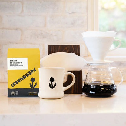 The Hario V60 Pro Bundle by Groundwork Coffee includes Bitches Brew coffee, a white mug, filters, and a pour-over maker on a light countertop.