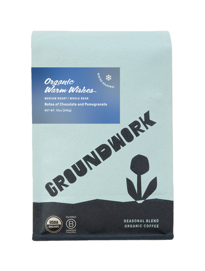 Groundwork Organic Warm Wishes winter seasonal coffee bag in light blue packaging with a blue label and tulip graphic. Text reads “Organic Warm Wishes,” “Medium Roast,” “Whole Bean,” “Notes of Chocolate and Pomegranate,” and “Net Wt 12oz (340g)” with USDA Organic and Certified B Corp seals shown at the bottom.