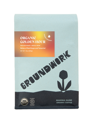 12oz bag of Groundwork Organic Golden Hour coffee. The packaging features a light blue upper section with a bold diagonal “GROUNDWORK” logo, black base, and stylized flower icon. A label reads “Organic Golden Hour – Medium Roast | Whole Bean – Notes of Red Grape and Tamarind,” and includes a “Summer Seasonal” mark. USDA Organic and Certified B Corporation logos appear at the bottom.