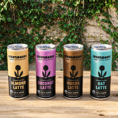 Four cans of Groundwork Coffee Organic Cold Brew Latte—Almond, Coconut, Mocha, and Oat—rest on a table with ivy and stone behind. Enjoy these delicious plant-based lattes.