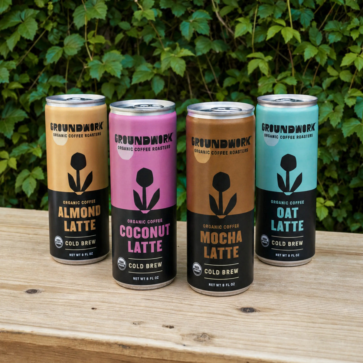 Four cans of Groundwork Coffee’s Organic Cold Brew Latte—Almond, Coconut, Mocha, and Oat—on wood with green leaves; each a plant-based latte with delicious flavor.