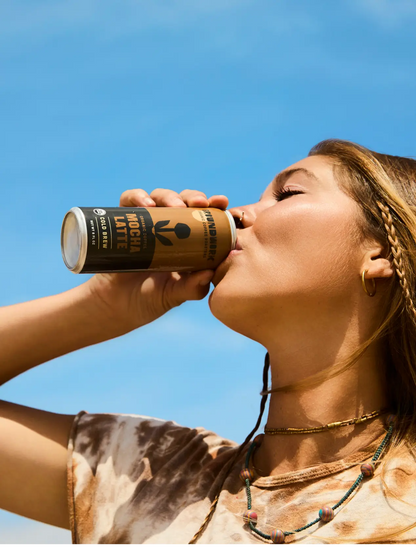 A woman with braided hair sips Groundwork Coffee’s Organic Cold Brew Latte from a can, wearing a tie-dye shirt and layered necklaces under a clear blue sky.