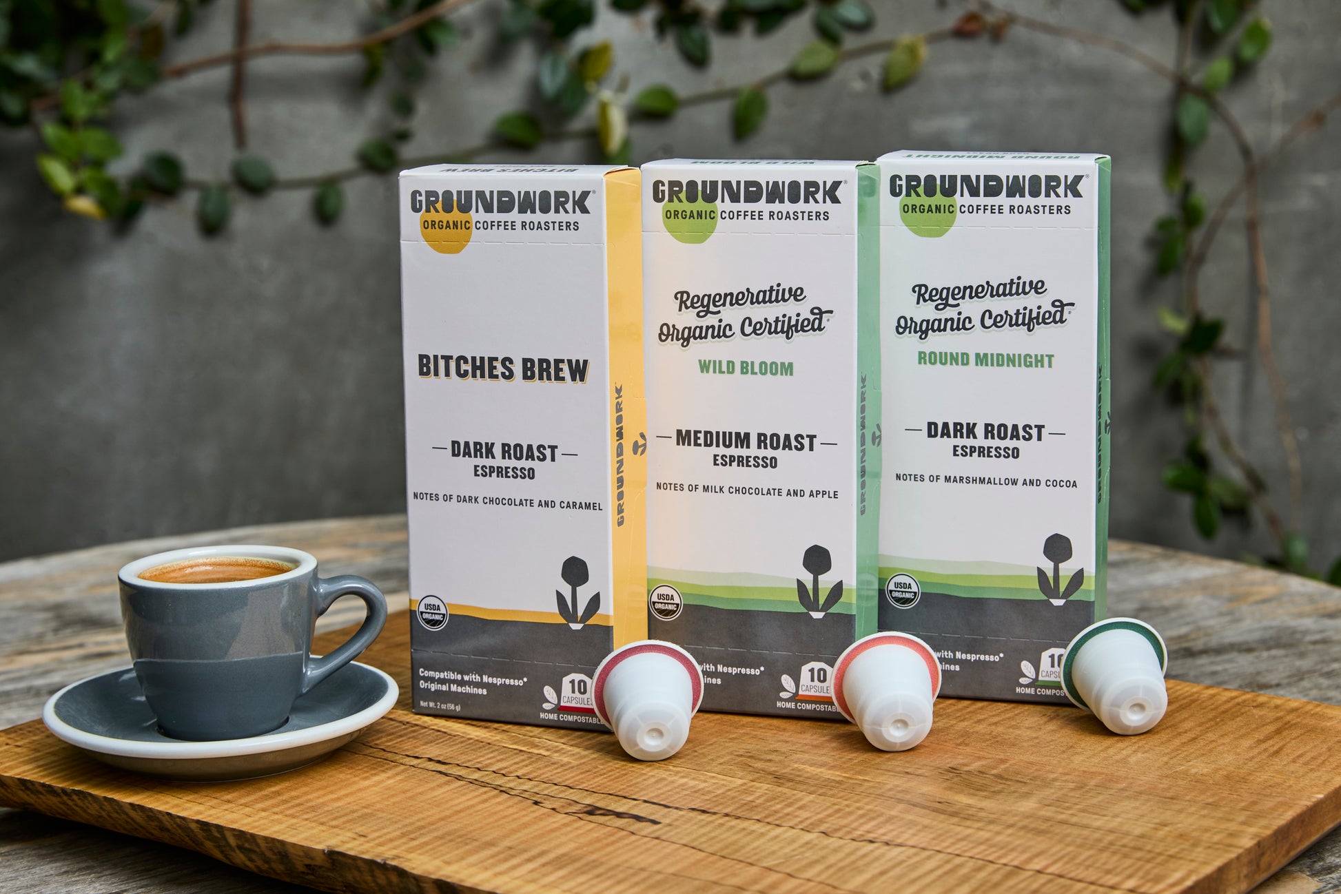 Three boxes of Groundwork Coffee Nespresso Variety Pack and a cup of espresso on a wooden tray, with greenery blurred in the background.