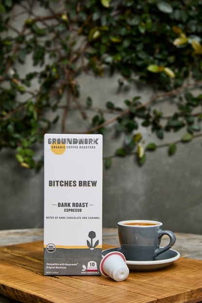 A box of Groundwork Coffee Organic Bitches Brew Nespresso pods sits on a wooden table next to an espresso cup, with green plants blurred in the background.