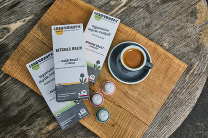 A cup of espresso on a saucer rests on a wooden table next to Groundwork Coffee's Nespresso Variety Pack and Groundwork regenerative organic coffee packages.