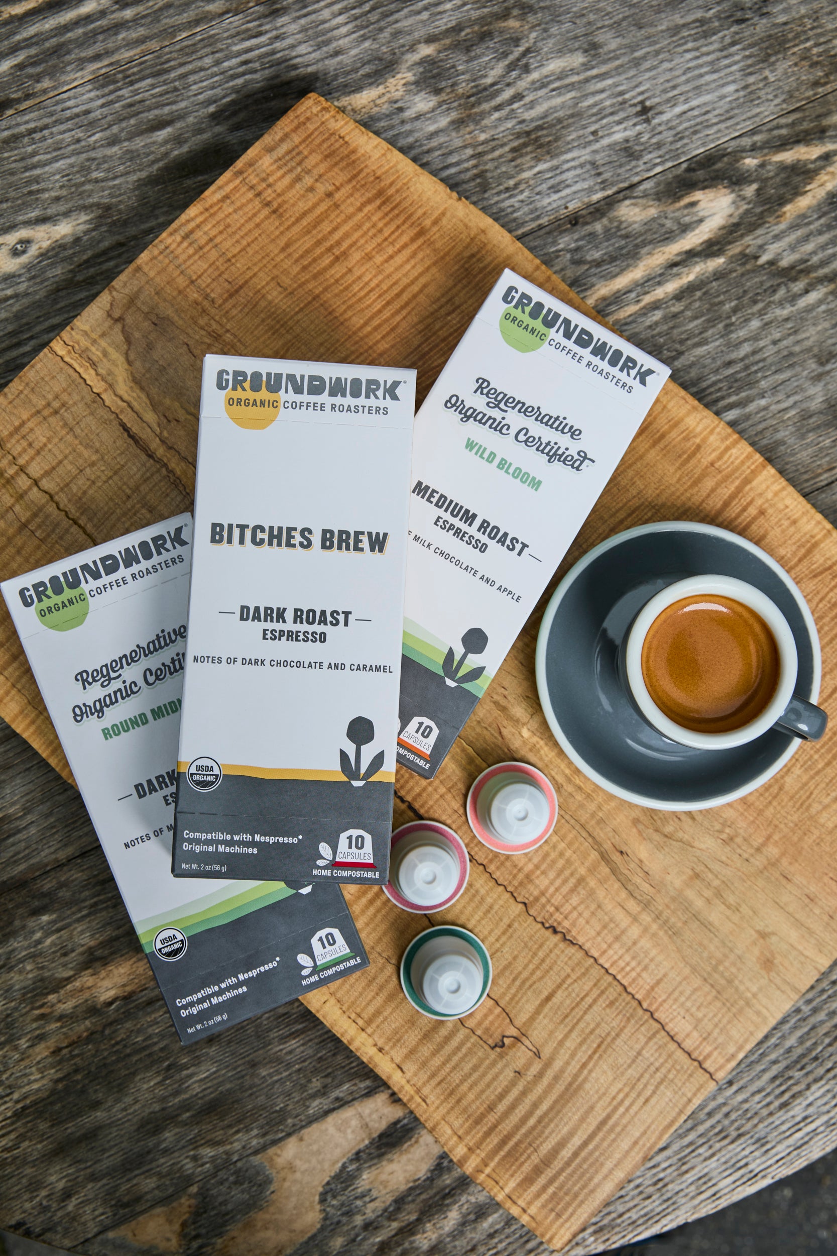 A cup of espresso sits on a wooden board next to three Groundwork Coffee Nespresso Variety Pack boxes and three colorful pods.
